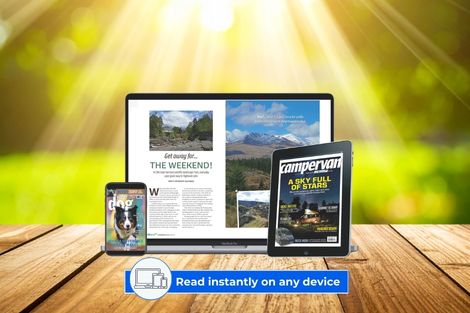 Laptop, tablet, and smartphone displaying the digital motorhomers magazine content on a wooden table outdoors with sun rays and greenery in the background