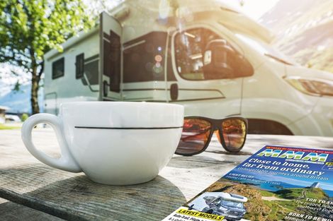 White coffee cup and sunglasses on a wooden picnic table beside an RV, with the motorhomers magazine in the foreground, set outdoors in a sunny campground.