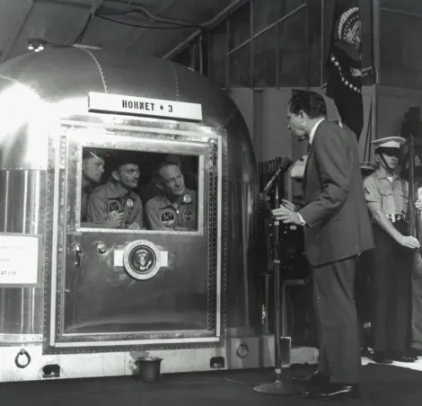 The Apollo 11 astronauts, left to right, Commander Neil A. Armstrong, Command Module Pilot Michael Collins and Lunar Module Pilot Edwin E. "Buzz" Aldrin Jr., inside the Mobile Quarantine Facility aboard the USS Hornet, listen to President Richard M. Nixon on July 24, 1969 as he welcomes them back to Earth and congratulates them.